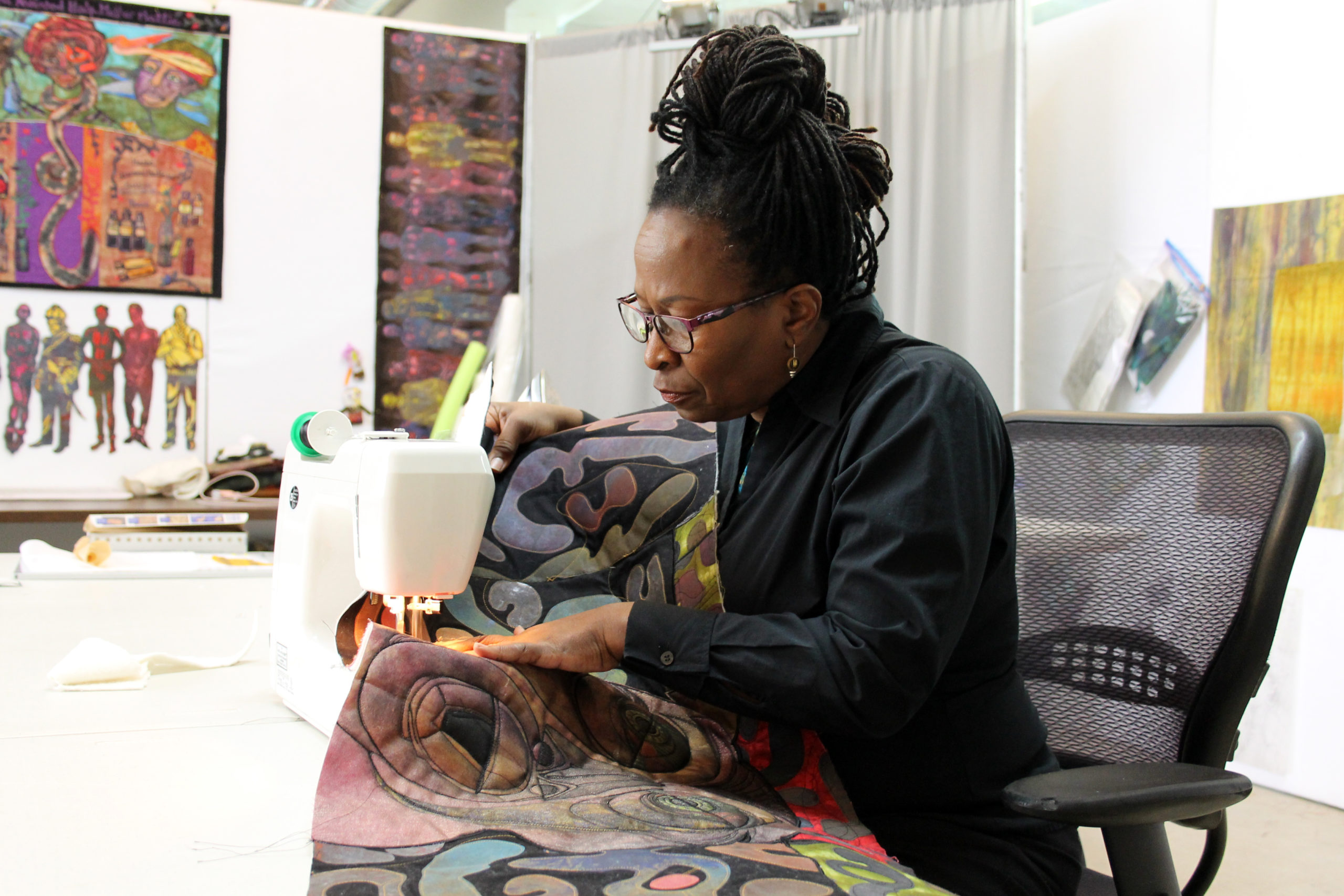 A black woman with dreadlocks piled on her head sewing part of a quilt on a sewing machine with her work surrounding her