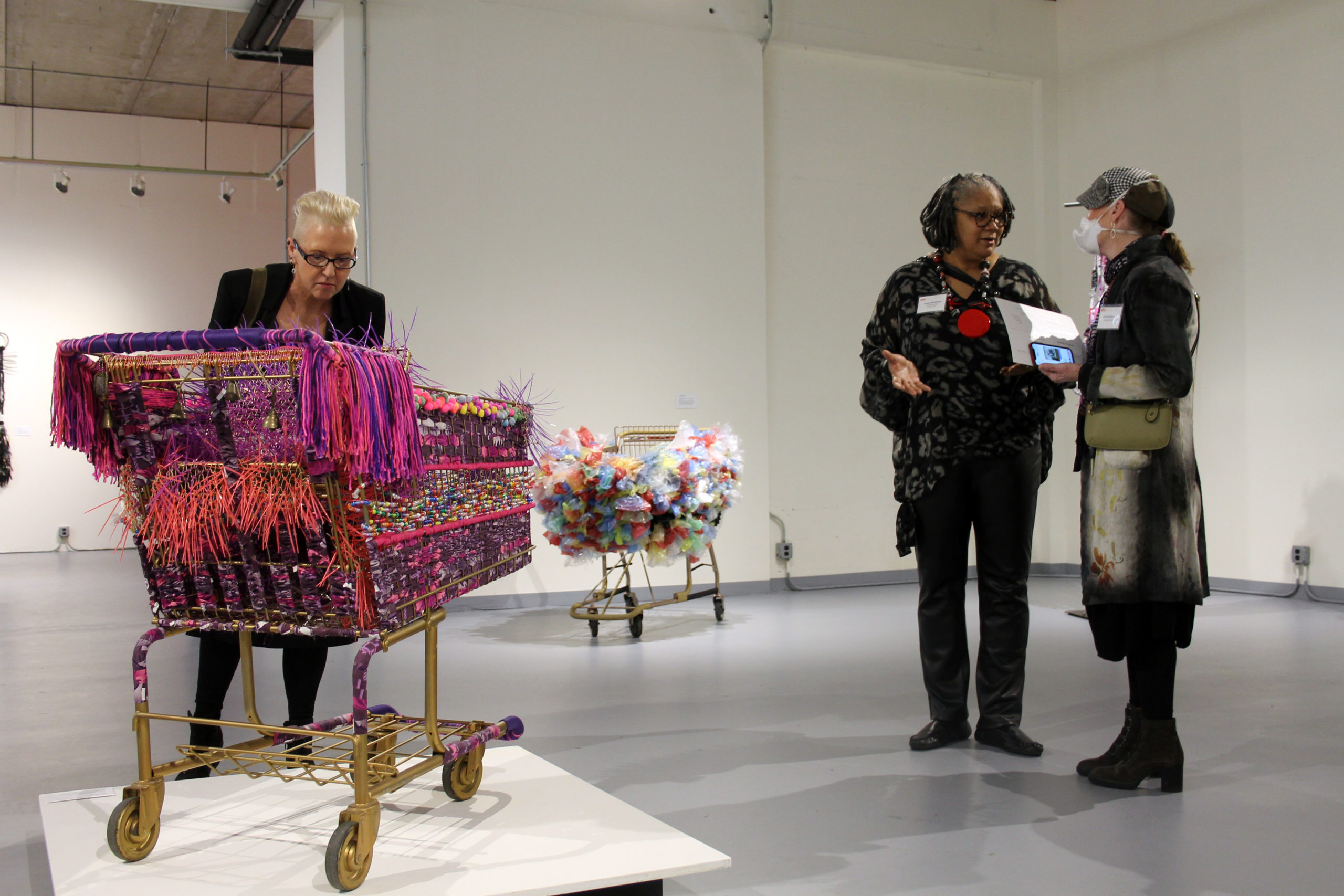 Two women, including artist Theda Sandiford, stand and talk on the right half of the image while another woman with very light hair and glasses closely examines a grocery cart festooned with ribbons, beads, and zip ties