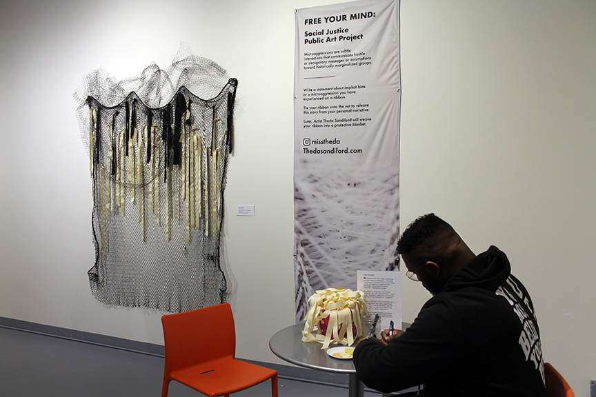 A man sits at a round table writing on a piece of ribbon that he will later attach to a fishing net that is hanging on a wall in the background