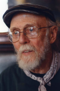 a head-and-shoulders shot of an elderly white man with a white beard and mustache wearing wire-rim glasses, a cap, and a bandana tied around his neck