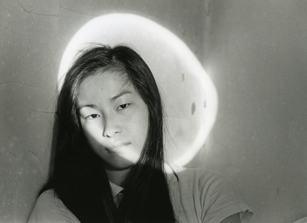 A black-haired woman of oriental descent looks at the camera with a circle of light reflected on her head and the wall behind her, looking like a halo