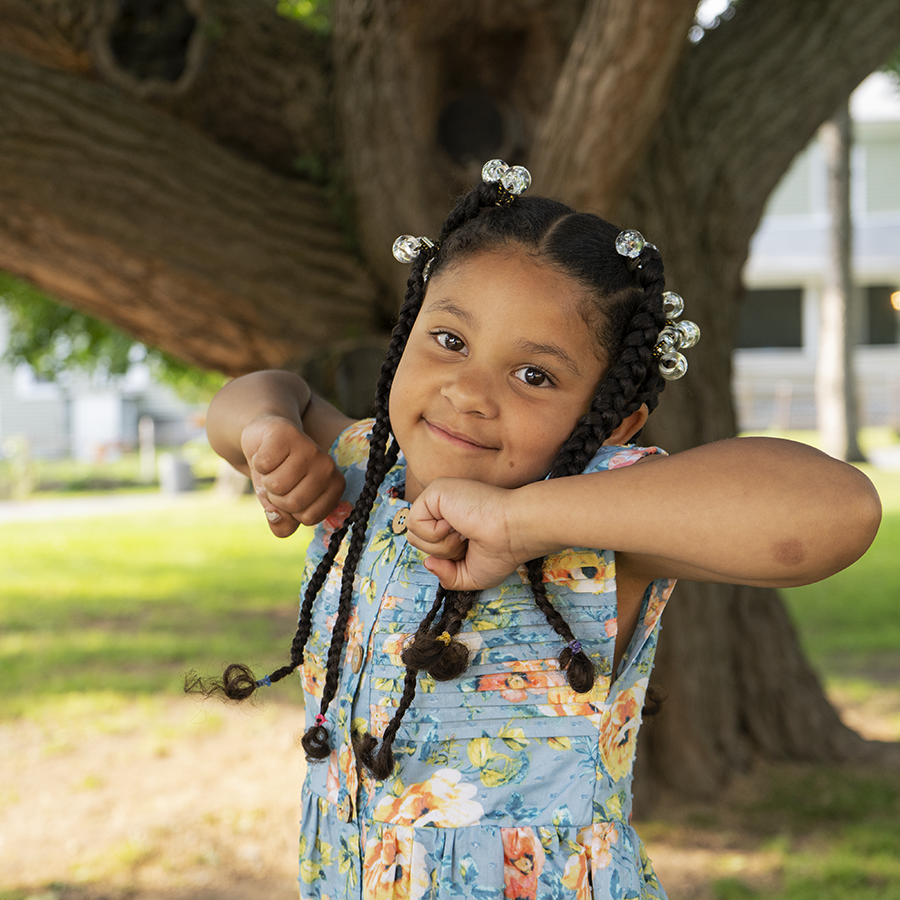 A young girl with a light brown complexion and several braids in her black hair is wearing a floral top and stands in front of an old tree.