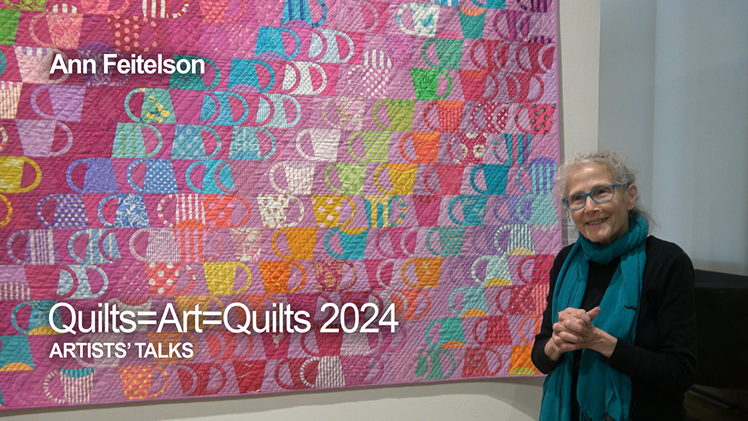 An older woman with white hair wearing black clothes and a bright blue scarf stands in front of her art quilt made up of dozens of red, pink, blue, yellow, and green coffee cups arranged in diagonal lines.