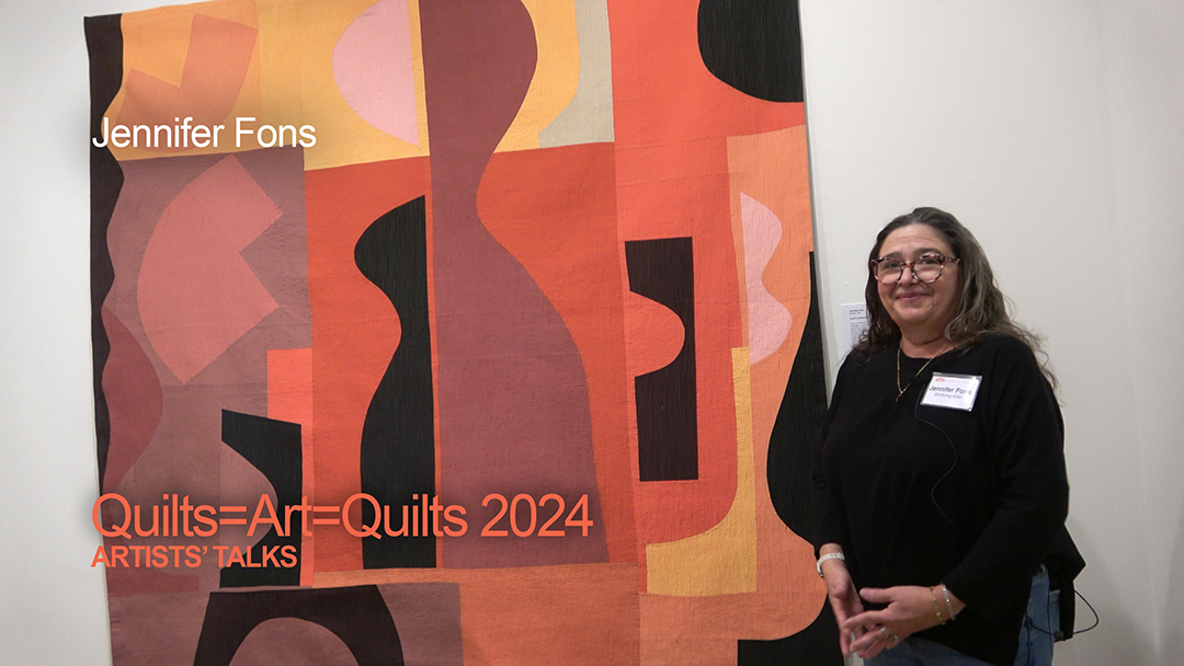 A white woman with dark hair wearing black clothing stands near an art quilt she made that eatures shades of maroon, orange, and black