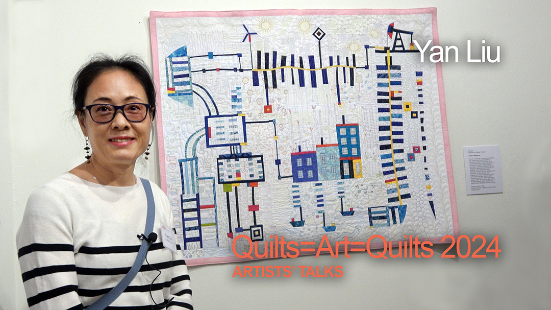 A Chinese woman with dark hair wearing glasses and a black and white striped sweater stands in front of her art quilt, which shows various forms of energy: Gas, oil, solar, and wind.