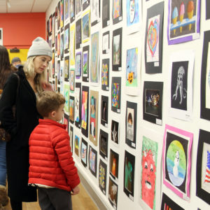 A boy wearing a red coat and his mother look at student artwork filling a wall from top to bottom in an art gallery