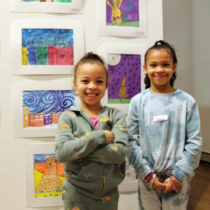 Two mixed-race young girls stand in front of student artwork, including a piece that the girl on the right made