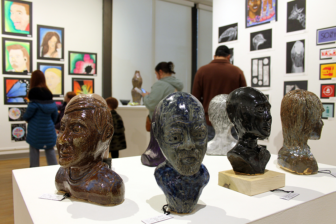 Several student sculptures of heads rest on a table while several adults and children look at student artwork in the background