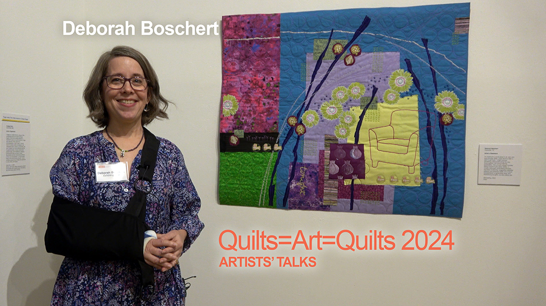 A white woman with blonde hair wearing a flowered shirt with her right arm in a sling stands in front of her colorful art quilt, which includes blue, red, yellow, green, and a stitched comfy arm chair, bunnies, and more.