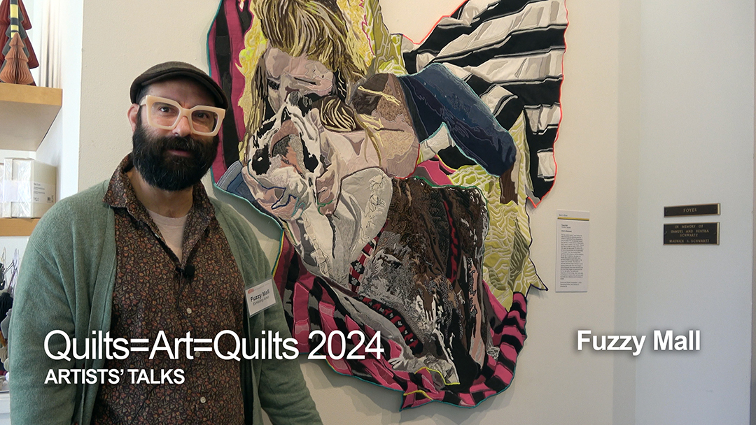 A white man with dark hair and a beard wearing a hat and a green sweater over a brown shirt stands in front of his art quilt, which shows a blonde woman's face and parts of a dog with material spread out like wings.