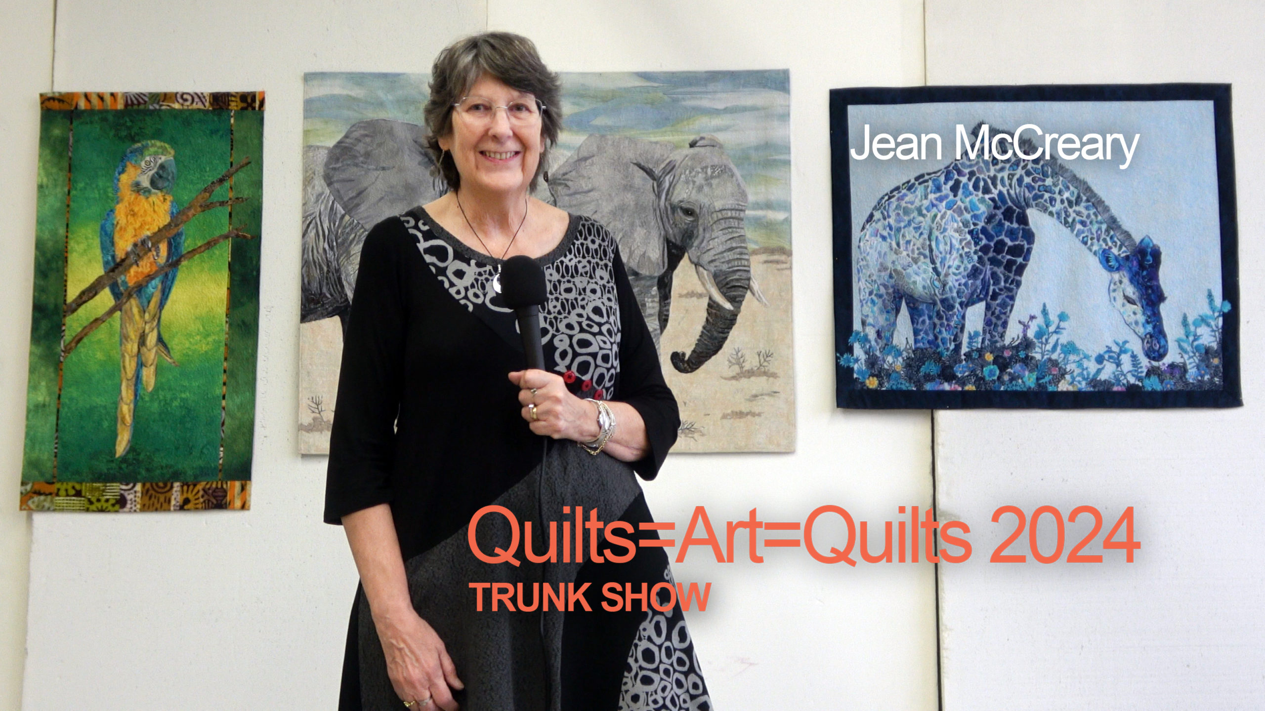 An older woman with short, graying hair wearing a black and gray top smiles as she stands in front of several of her art quilts featuring a macaw, elephants and a giraffe.