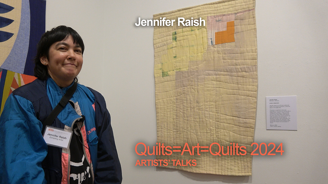 A woman with short dark hair wearing a blue and black top stands by her art quilt, which is mostly pale yellow and beige with a section of an orangey-red.