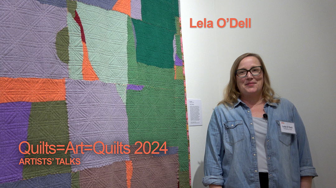 A white woman with long blonde hair wearing a jean's shirt stands in front of herart quilt, which is made of gray, green, orage and purple recycled cashmere.